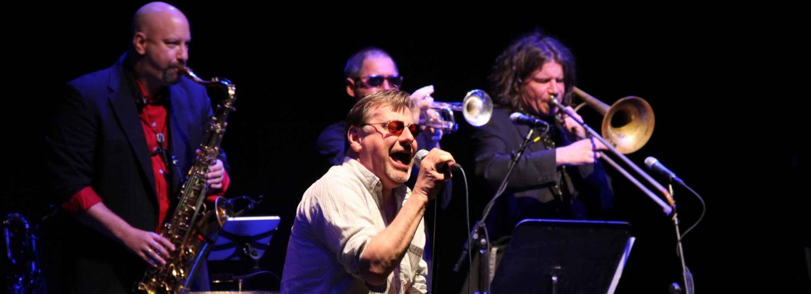 Southside Johnny singing into a microphone with horn players in the background