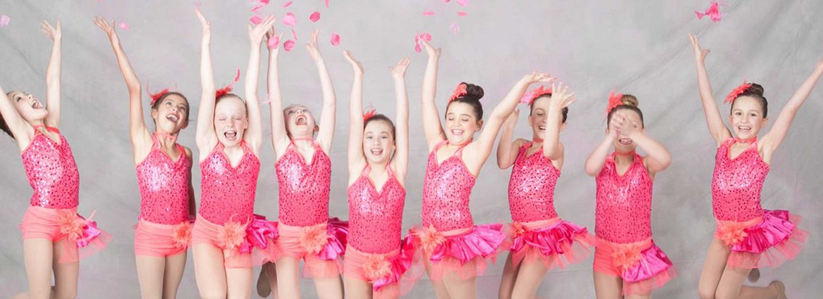 Nine young female dancers dressed in their dance outfit and tutu's throwing pink rose petals while posing for a picture.